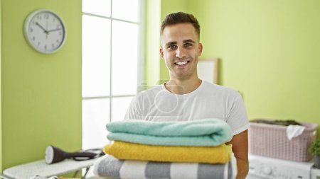 Téléchargez les photos : Beau jeune homme hispanique souriant dans une buanderie bien éclairée, tenant des serviettes pliées avec une machine à laver en arrière-plan - en image libre de droit