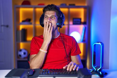 Foto de Joven hispano jugando videojuegos aburrido bostezando cansado cubriendo la boca con la mano. inquietud y somnolencia. - Imagen libre de derechos