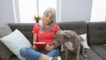 Foto de Relajada mujer de mediana edad, una bibliófila de pelo gris, acurrucada con su leal perro, cómodamente sentada en su sofá de casa, disfrutando del ocio de leer un libro fascinante en su acogedora sala de estar. - Imagen libre de derechos