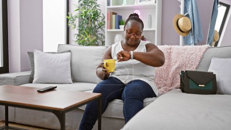 Téléchargez les photos : Femme afro-américaine détendue savourant l'expresso du matin, confortablement assise sur le canapé de la maison, regardant la montre avec une expression sérieuse - en image libre de droit