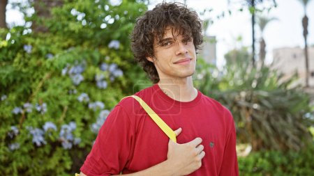 Photo for Handsome young man with curly hair smiling outdoors in a park wearing a red shirt and yellow strap. - Royalty Free Image