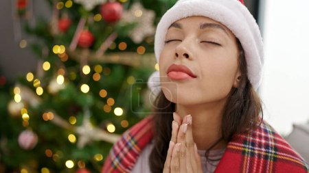 Photo for Young beautiful hispanic woman praying sitting by christmas tree at home - Royalty Free Image