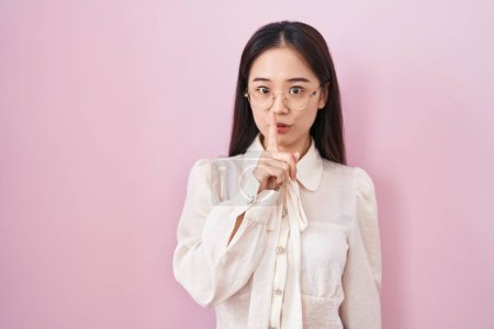 Téléchargez les photos : Jeune femme chinoise debout sur fond rose demandant à être tranquille avec le doigt sur les lèvres. silence et concept secret. - en image libre de droit