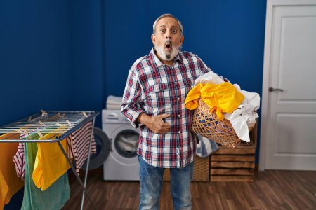 Photo for Amazed senior man, mouth wide open in shock, holding a laundry basket of clothes in the laundry room - a picture of astonish and fear, a portrait of funny disbelief! - Royalty Free Image