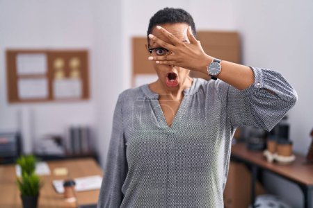 Photo for African american woman working at the office wearing glasses peeking in shock covering face and eyes with hand, looking through fingers with embarrassed expression. - Royalty Free Image