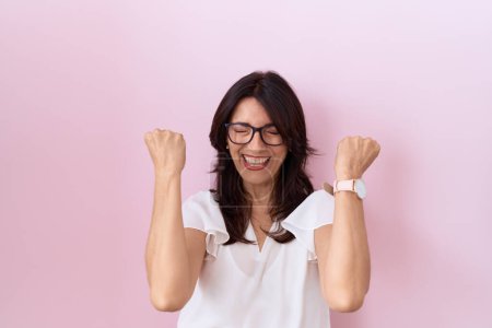 Foto de Mujer hispana de mediana edad con camiseta blanca casual y gafas celebrando sorprendida y sorprendida por el éxito con los brazos levantados y los ojos cerrados. concepto ganador. - Imagen libre de derechos