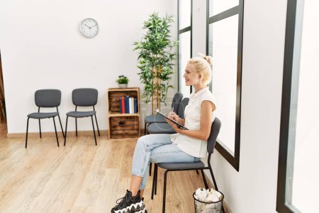 Téléchargez les photos : Jeune femme blonde écrit sur le presse-papiers assis sur la chaise dans la salle d'attente - en image libre de droit