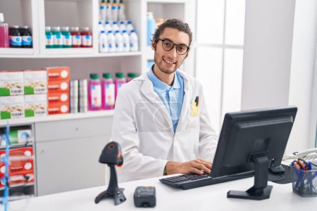 Téléchargez les photos : Jeune homme hispanique pharmacien souriant confiant en utilisant l'ordinateur à la pharmacie - en image libre de droit