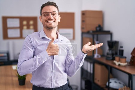 Téléchargez les photos : Jeune homme hispanique au bureau montrant la main de la paume et faisant un geste correct avec les pouces levés, souriant heureux et joyeux - en image libre de droit