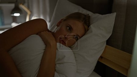 Téléchargez les photos : Jeune femme blonde allongée sur le lit avec un visage triste étreignant la couverture dans la chambre - en image libre de droit