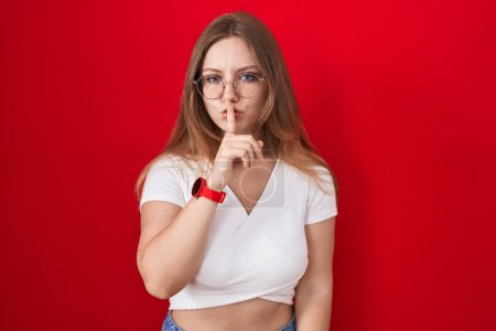 Téléchargez les photos : Jeune femme caucasienne debout sur fond rouge demandant d'être calme avec le doigt sur les lèvres. silence et concept secret. - en image libre de droit