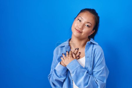 Photo for Asian young woman standing over blue background smiling with hands on chest with closed eyes and grateful gesture on face. health concept. - Royalty Free Image