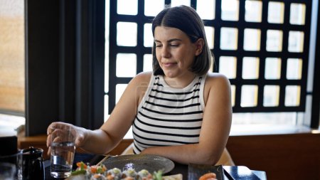 Junge schöne hispanische Frau isst Sushi und trinkt Wasser im Restaurant