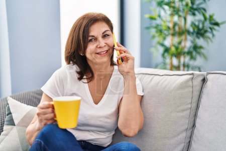 Foto de Middle age woman talking on smartphone drinking coffee at home - Imagen libre de derechos
