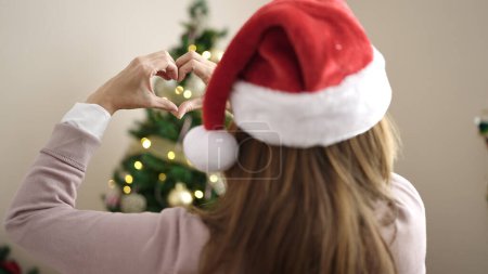 Foto de Joven mujer hispana hermosa haciendo gesto de corazón de pie junto al árbol de Navidad en casa - Imagen libre de derechos