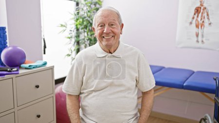 Téléchargez les photos : Homme âgé joyeux avec les cheveux blancs, assis avec confiance sur la chaise, souriant tout en recevant un traitement de physiothérapie à la clinique de réadaptation - en image libre de droit