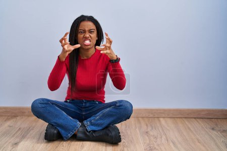 Photo for Young african american with braids sitting on the floor at home shouting frustrated with rage, hands trying to strangle, yelling mad - Royalty Free Image