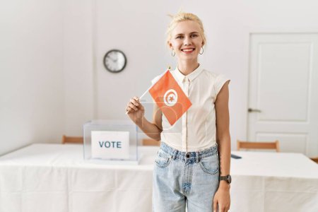Photo for Young blonde woman holding tunisia flag smiling at electoral college - Royalty Free Image