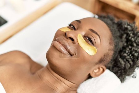 Téléchargez les photos : Femme afro-américaine allongée sur une table de massage ayant un traitement de masque oculaire au salon de beauté - en image libre de droit