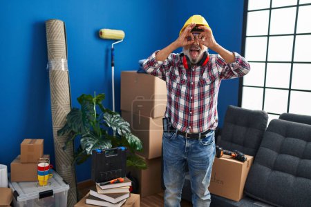 Photo for Cheeky senior man in hardhat and safety glasses, pulling a fun, approving face, sticking tongue out and looking through the 'ok' hand gesture like binoculars at his new home - Royalty Free Image
