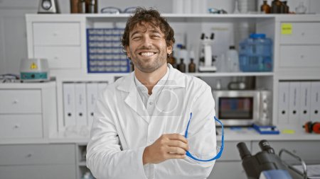 Téléchargez les photos : Homme hispanique souriant avec barbe portant un blouse de laboratoire dans un environnement de laboratoire moderne - en image libre de droit