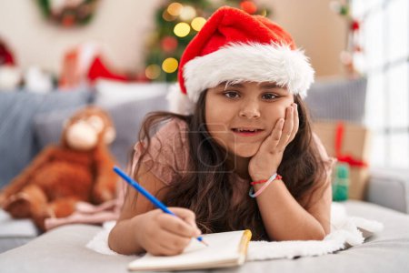 Photo for Plus size hispanic girl writing on notebook lying on sofa by christmas tree at home - Royalty Free Image