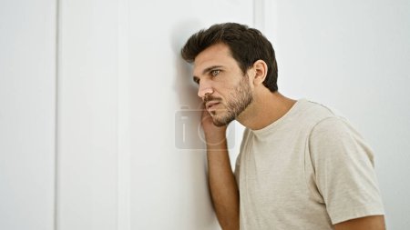 Photo for Hispanic young man with a beard casually leaning on a white interior door at home, looking pensive. - Royalty Free Image