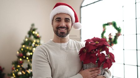 Téléchargez les photos : Jeune homme hispanique tenant une plante debout près de l'arbre de Noël à la maison - en image libre de droit