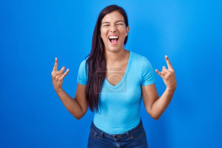 Foto de Mujer hispana joven de pie sobre fondo azul gritando con expresión loca haciendo símbolo de roca con las manos arriba. Estrella de la música. concepto pesado. - Imagen libre de derechos