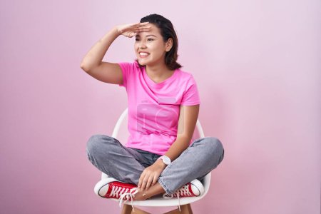 Téléchargez les photos : Jeune femme hispanique assise sur une chaise sur fond rose très heureuse et souriante regardant loin avec la main sur la tête. concept de recherche. - en image libre de droit