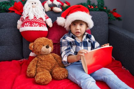Photo for Adorable hispanic boy reading book sitting on sofa by christmas decoration at home - Royalty Free Image