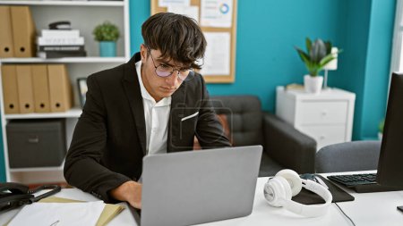 Foto de Adolescente hispano serio y joven que adopta la tecnología y trabaja como profesional de negocios en la oficina. sentado en su escritorio, investido de éxito, se sumerge en el trabajo en su computadora portátil - Imagen libre de derechos