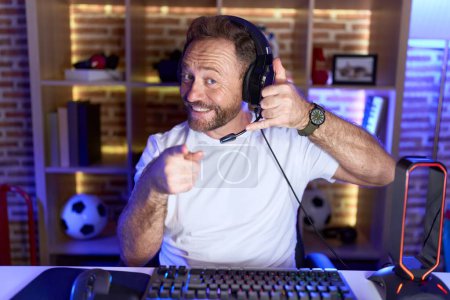 Foto de Hombre de mediana edad con barba jugando videojuegos con auriculares sonriendo hablando por teléfono y señalándote. Llámame.. - Imagen libre de derechos