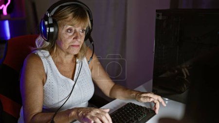 Photo for Beautiful middle age blonde woman streamer seriously immersed, wearing headphones, playing video game. indoor night gaming room streaming with advanced tech gadget. - Royalty Free Image