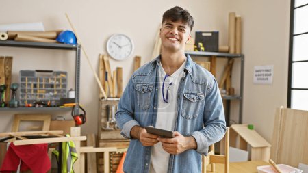 Photo for Smiling young hispanic man, a professional carpenter, engrossed in his carpentry work indoors, skillfully using a touchpad in his well-furnished workshop - Royalty Free Image