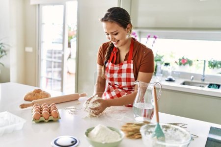 Foto de Joven hermosa mujer hispana sonriendo confiada amasando masa de pan con las manos en la cocina - Imagen libre de derechos