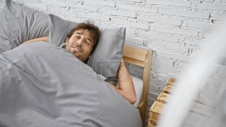 Photo for Handsome hispanic man with beard and green eyes enjoys cool air from fan in a cozy bedroom setting. - Royalty Free Image