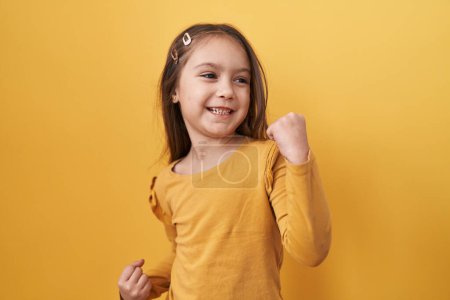 Photo for Adorable hispanic girl joyfully standing, arms raised in victorious celebration of success on a yellow isolated background, eyes softly closed, yes! - Royalty Free Image
