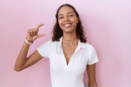 Téléchargez les photos : Jeune femme hispanique portant t-shirt blanc décontracté geste souriant et confiant avec la main faisant signe de petite taille avec les doigts à la recherche et la caméra. concept de mesure. - en image libre de droit