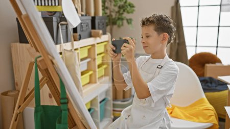 Foto de Adorable chico rubio artista, sonriendo mientras toma una foto en un estudio de arte, listo para dibujar su obra maestra sobre lienzo - Imagen libre de derechos
