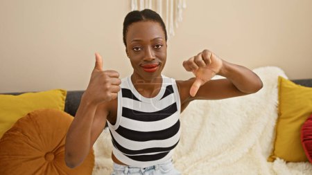 Photo for At home on the sofa, a confident african american woman gives a thumb up gesture, then flashes a thumb down. fun and expressive indoors! - Royalty Free Image
