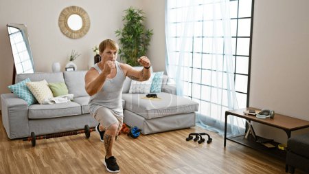 Photo for A young man with blond hair and tattoos lunging in his living room, surrounded by workout equipment. - Royalty Free Image