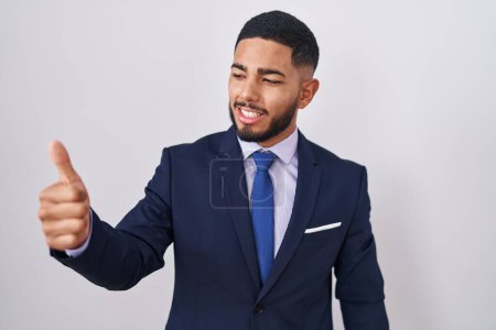 Téléchargez les photos : Jeune homme hispanique portant un costume d'affaires et cravate fière, souriant en faisant les pouces vers le haut geste sur le côté - en image libre de droit