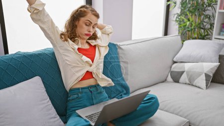 Photo for Exhausted young woman relaxing, stretching tired arms back on sofa, using laptop indoors at home - Royalty Free Image