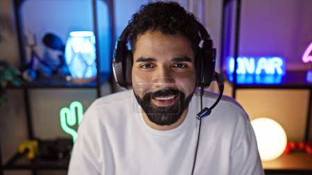 Photo for A handsome young hispanic man with a beard wearing headphones smiles in a neon-lit gaming room at night. - Royalty Free Image
