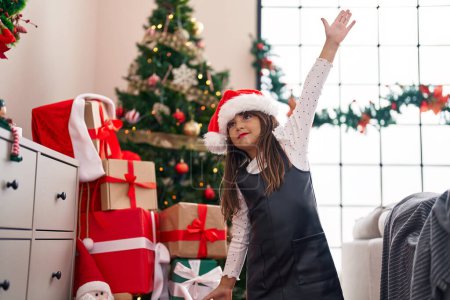 Foto de Adorable chica hispana sonriendo confiada de pie junto al árbol de Navidad en casa - Imagen libre de derechos