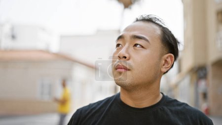 Téléchargez les photos : Cool et détendu, un jeune et beau Chinois qui jette une expression sérieuse, regardant du côté d'une rue urbaine sous le soleil - en image libre de droit