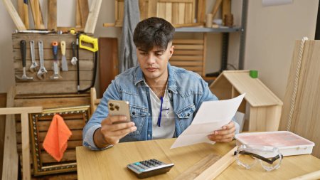 Foto de Guapo joven hispano inmerso en su negocio de carpintería, cautivado por un documento en su teléfono inteligente en medio de un taller interior lleno de madera. - Imagen libre de derechos
