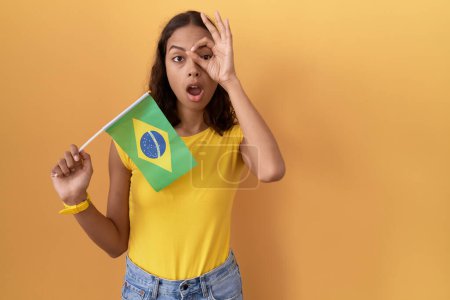 Foto de Mujer hispana joven sosteniendo la bandera de Brasil haciendo un gesto bien sorprendido con la cara sorprendida, los ojos mirando a través de los dedos. expresión incrédula. - Imagen libre de derechos
