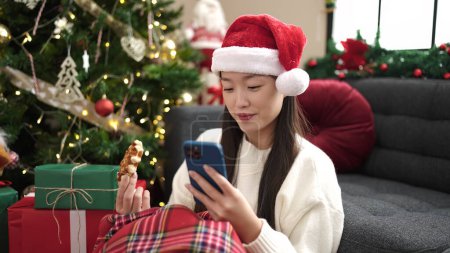 Foto de Joven mujer china desayunando usando smartphone sentado junto al árbol de Navidad en casa - Imagen libre de derechos